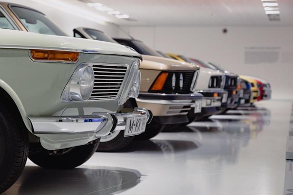 A row of vintage cars, mainly BMWs, is parked inside a bright, modern showroom. The image focuses on the front grills and bumpers, showcasing classic designs and glossy finishes. | MONEY6X