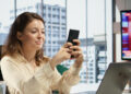 A woman sitting at a desk in a modern office smiles while using her smartphone. A laptop and stationery are on the desk. Large windows show city buildings, and a TV displays news in the background. | MONEY6X