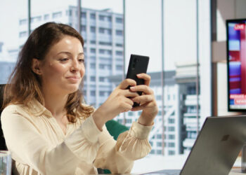 A woman sitting at a desk in a modern office smiles while using her smartphone. A laptop and stationery are on the desk. Large windows show city buildings, and a TV displays news in the background. | MONEY6X