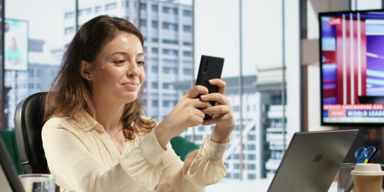 A woman sitting at a desk in a modern office smiles while using her smartphone. A laptop and stationery are on the desk. Large windows show city buildings, and a TV displays news in the background. | MONEY6X