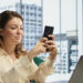 A woman sitting at a desk in a modern office smiles while using her smartphone. A laptop and stationery are on the desk. Large windows show city buildings, and a TV displays news in the background. | MONEY6X
