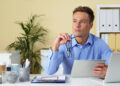A man in a blue shirt sits at a desk holding a tablet and eyeglasses, looking thoughtful. Office supplies and folders are on the desk; a plant and shelves are in the background. | MONEY6X