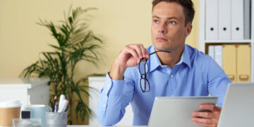 A man in a blue shirt sits at a desk holding a tablet and eyeglasses, looking thoughtful. Office supplies and folders are on the desk; a plant and shelves are in the background. | MONEY6X
