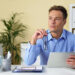 A man in a blue shirt sits at a desk holding a tablet and eyeglasses, looking thoughtful. Office supplies and folders are on the desk; a plant and shelves are in the background. | MONEY6X