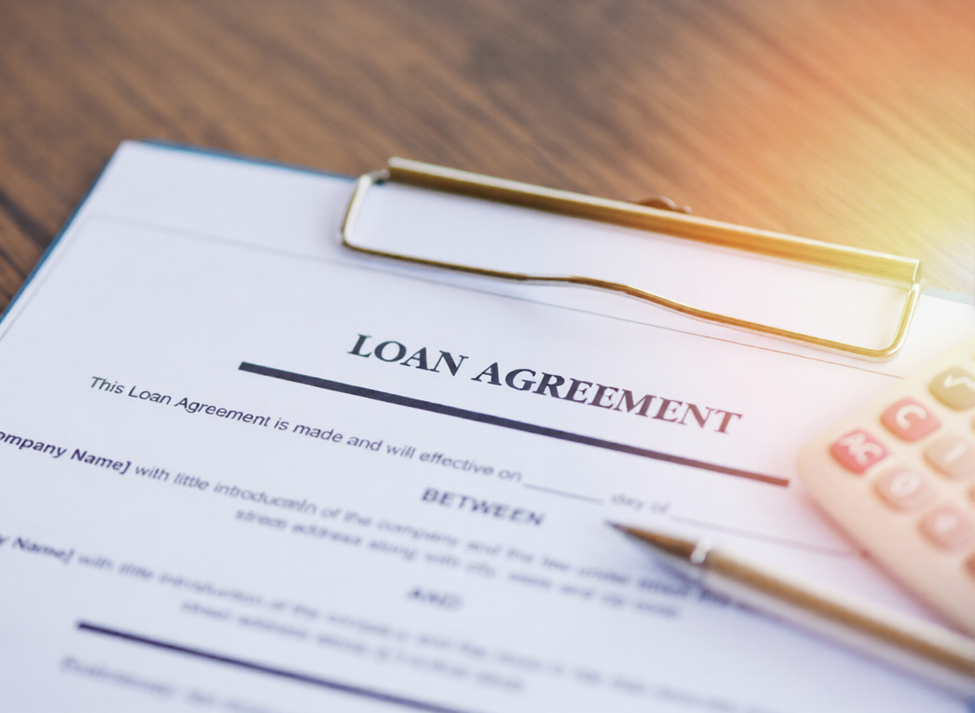 A close-up of a loan agreement document on a clipboard, with a pen and a calculator placed nearby on a wooden desk under bright light. | MONEY6X