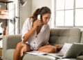 A woman with glasses sits on a couch, holding a smartphone and looking at papers and a laptop on the table. She appears focused, surrounded by documents and working in a bright room with a large window. | MONEY6X