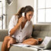 A woman with glasses sits on a couch, holding a smartphone and looking at papers and a laptop on the table. She appears focused, surrounded by documents and working in a bright room with a large window. | MONEY6X