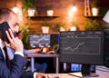 A man in a suit talks on the phone while looking at financial graphs on dual computer monitors in a modern office with plants on wooden shelves in the background. | MONEY6X