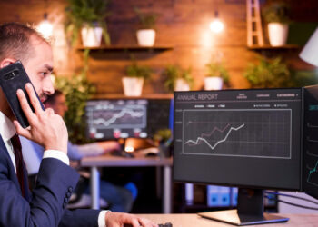 A man in a suit talks on the phone while looking at financial graphs on dual computer monitors in a modern office with plants on wooden shelves in the background. | MONEY6X