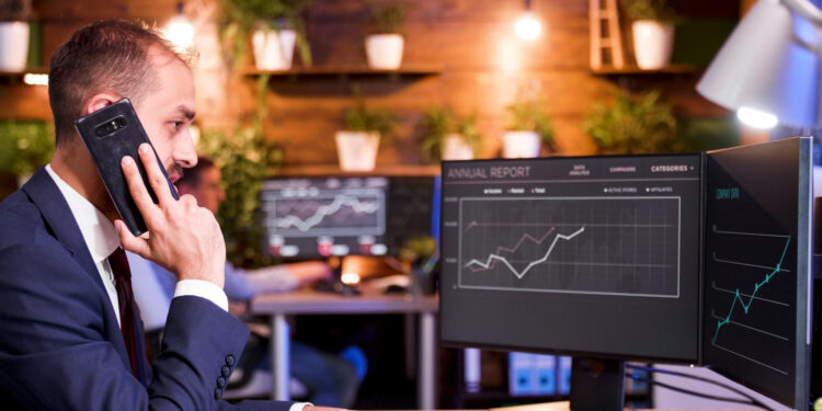 A man in a suit talks on the phone while looking at financial graphs on dual computer monitors in a modern office with plants on wooden shelves in the background. | MONEY6X