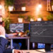A man in a suit talks on the phone while looking at financial graphs on dual computer monitors in a modern office with plants on wooden shelves in the background. | MONEY6X