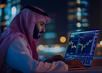 A man in traditional Middle Eastern attire analyzes colorful financial charts on a laptop at night, with blurred city lights in the background. | MONEY6X