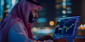 A man in traditional Middle Eastern attire analyzes colorful financial charts on a laptop at night, with blurred city lights in the background. | MONEY6X