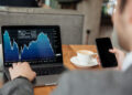 A person in a suit looks at a laptop displaying a downward-trending stock chart while holding a smartphone, with a cup of coffee on the table beside them. | MONEY6X