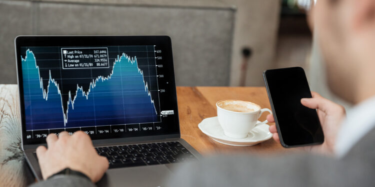 A person in a suit looks at a laptop displaying a downward-trending stock chart while holding a smartphone, with a cup of coffee on the table beside them. | MONEY6X