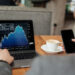 A person in a suit looks at a laptop displaying a downward-trending stock chart while holding a smartphone, with a cup of coffee on the table beside them. | MONEY6X