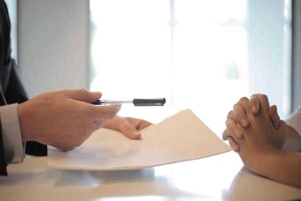 A person in a suit hands a pen and documents to another person with clasped hands, suggesting a signing or agreement at a desk with a bright window in the background. | MONEY6X