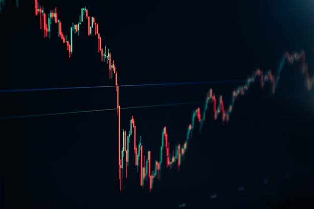 A digital display of a financial stock chart shows a sharp downward trend followed by a gradual upward movement, with red and green candlesticks indicating price changes on a dark background. | MONEY6X