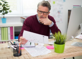A man with gray hair and glasses sits at a desk, looking thoughtfully at a sheet of paper with graphs. He is surrounded by office supplies, a plant, and a computer in a bright office space. | MONEY6X