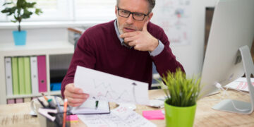 A man with gray hair and glasses sits at a desk, looking thoughtfully at a sheet of paper with graphs. He is surrounded by office supplies, a plant, and a computer in a bright office space. | MONEY6X