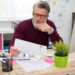 A man with gray hair and glasses sits at a desk, looking thoughtfully at a sheet of paper with graphs. He is surrounded by office supplies, a plant, and a computer in a bright office space. | MONEY6X