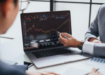 Two people in business attire discuss a rising stock chart displayed on a laptop screen, with one person pointing at the graph using a pen. Financial documents are visible on the table. | MONEY6X