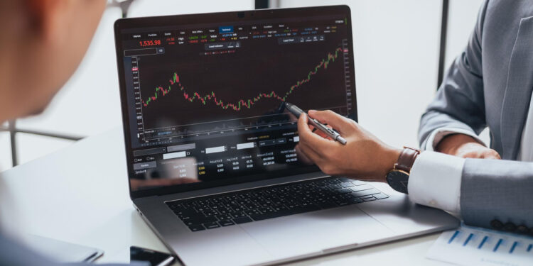 Two people in business attire discuss a rising stock chart displayed on a laptop screen, with one person pointing at the graph using a pen. Financial documents are visible on the table. | MONEY6X