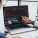 Two people in business attire discuss a rising stock chart displayed on a laptop screen, with one person pointing at the graph using a pen. Financial documents are visible on the table. | MONEY6X