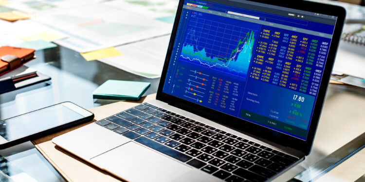 A laptop displaying colorful stock market charts and financial data sits on a glass desk, surrounded by papers, sticky notes, and a smartphone. | MONEY6X