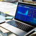 A laptop displaying colorful stock market charts and financial data sits on a glass desk, surrounded by papers, sticky notes, and a smartphone. | MONEY6X