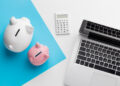A white and a pink piggy bank, a white calculator, and a silver laptop on a white and blue background, symbolizing savings and financial planning. | MONEY6X