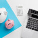 A white and a pink piggy bank, a white calculator, and a silver laptop on a white and blue background, symbolizing savings and financial planning. | MONEY6X