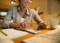 A woman smiles while using a smartphone and holding a credit card at a desk with a notebook, pencil, keyboard, and coffee mug, suggesting online shopping or banking. | MONEY6X