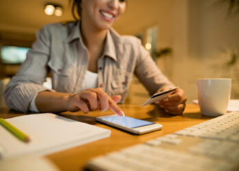 A woman smiles while using a smartphone and holding a credit card at a desk with a notebook, pencil, keyboard, and coffee mug, suggesting online shopping or banking. | MONEY6X
