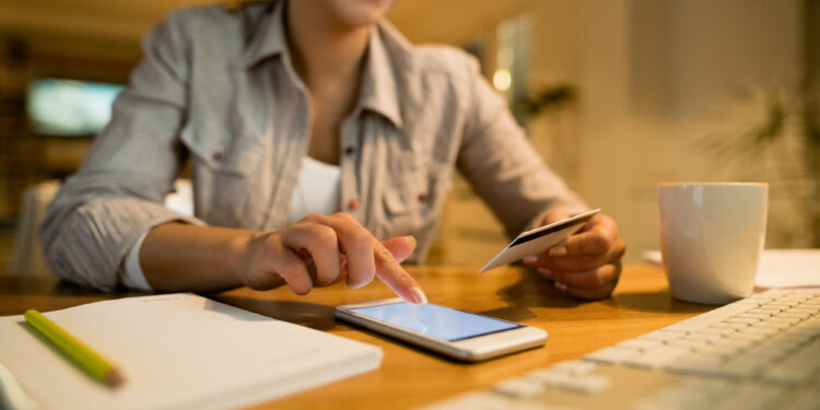 A woman smiles while using a smartphone and holding a credit card at a desk with a notebook, pencil, keyboard, and coffee mug, suggesting online shopping or banking. | MONEY6X