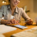 A woman smiles while using a smartphone and holding a credit card at a desk with a notebook, pencil, keyboard, and coffee mug, suggesting online shopping or banking. | MONEY6X