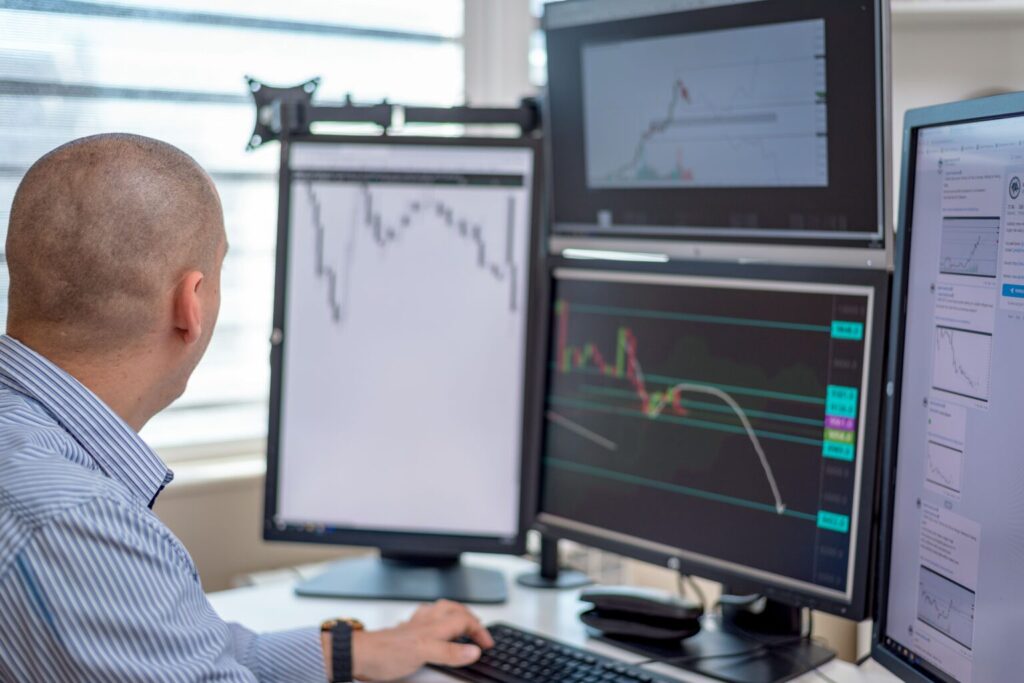 A person sits at a desk analyzing multiple computer monitors displaying stock charts and financial data, focusing intently on the screen in front of them. | MONEY6X