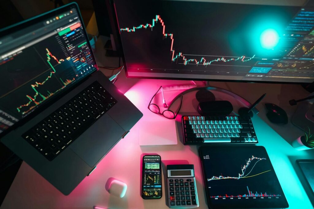 A desk with a laptop, monitor, keyboard, tablet, smartphone, calculator, glasses, and AirPods, all displaying financial charts and data, illuminated by colorful LED lights. | MONEY6X
