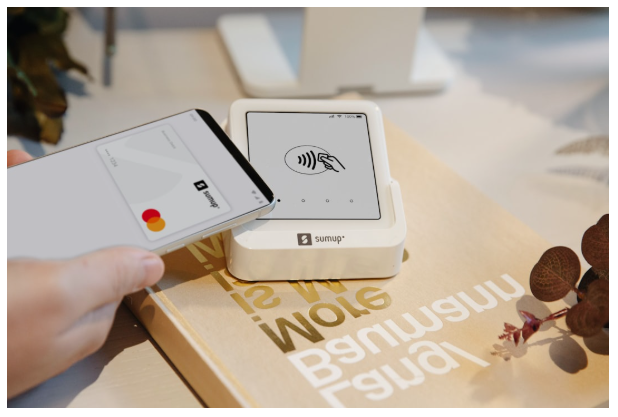 A person holds a smartphone near a SumUp card reader, ready to make a contactless payment. The device displays a contactless symbol and a hand holding a card. | MONEY6X