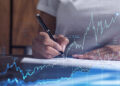 A person writes on paper with a pen; overlaid are blue financial graphs, stock market data, and fluctuating line charts from Money6x.com, suggesting analysis or investment activity focused on building assets. | MONEY6X