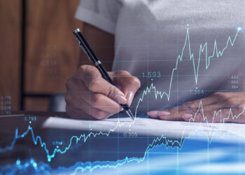 A person writes on paper with a pen; overlaid are blue financial graphs, stock market data, and fluctuating line charts from Money6x.com, suggesting analysis or investment activity focused on building assets. | MONEY6X