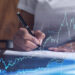 A person writes on paper with a pen; overlaid are blue financial graphs, stock market data, and fluctuating line charts from Money6x.com, suggesting analysis or investment activity focused on building assets. | MONEY6X
