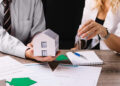 Two people sit at a table with mortgage documents; one holds a model house, while the other hands over house keys, symbolizing a real estate or home buying transaction. | MONEY6X