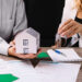 Two people sit at a table with mortgage documents; one holds a model house, while the other hands over house keys, symbolizing a real estate or home buying transaction. | MONEY6X