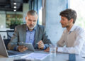Two men in business attire sit at a table with a laptop, tablet, glass of water, and documents, discussing something while one man points at the tablet screen. | MONEY6X