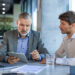 Two men in business attire sit at a table with a laptop, tablet, glass of water, and documents, discussing something while one man points at the tablet screen. | MONEY6X