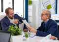Two men in business suits discuss documents at an office desk with laptops, papers, and potted plants, appearing focused and engaged in conversation. | MONEY6X