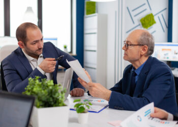 Two men in business suits discuss documents at an office desk with laptops, papers, and potted plants, appearing focused and engaged in conversation. | MONEY6X