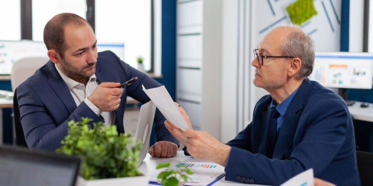 Two men in business suits discuss documents at an office desk with laptops, papers, and potted plants, appearing focused and engaged in conversation. | MONEY6X