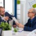 Two men in business suits discuss documents at an office desk with laptops, papers, and potted plants, appearing focused and engaged in conversation. | MONEY6X